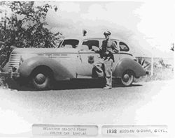 Black and white photo of a police car with an officer standing in front of it